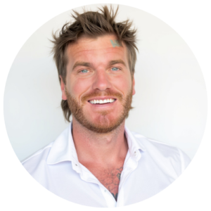 Atmos Healthcare Smiling man with tousled hair and a beard, wearing a white shirt, standing against a plain white background.